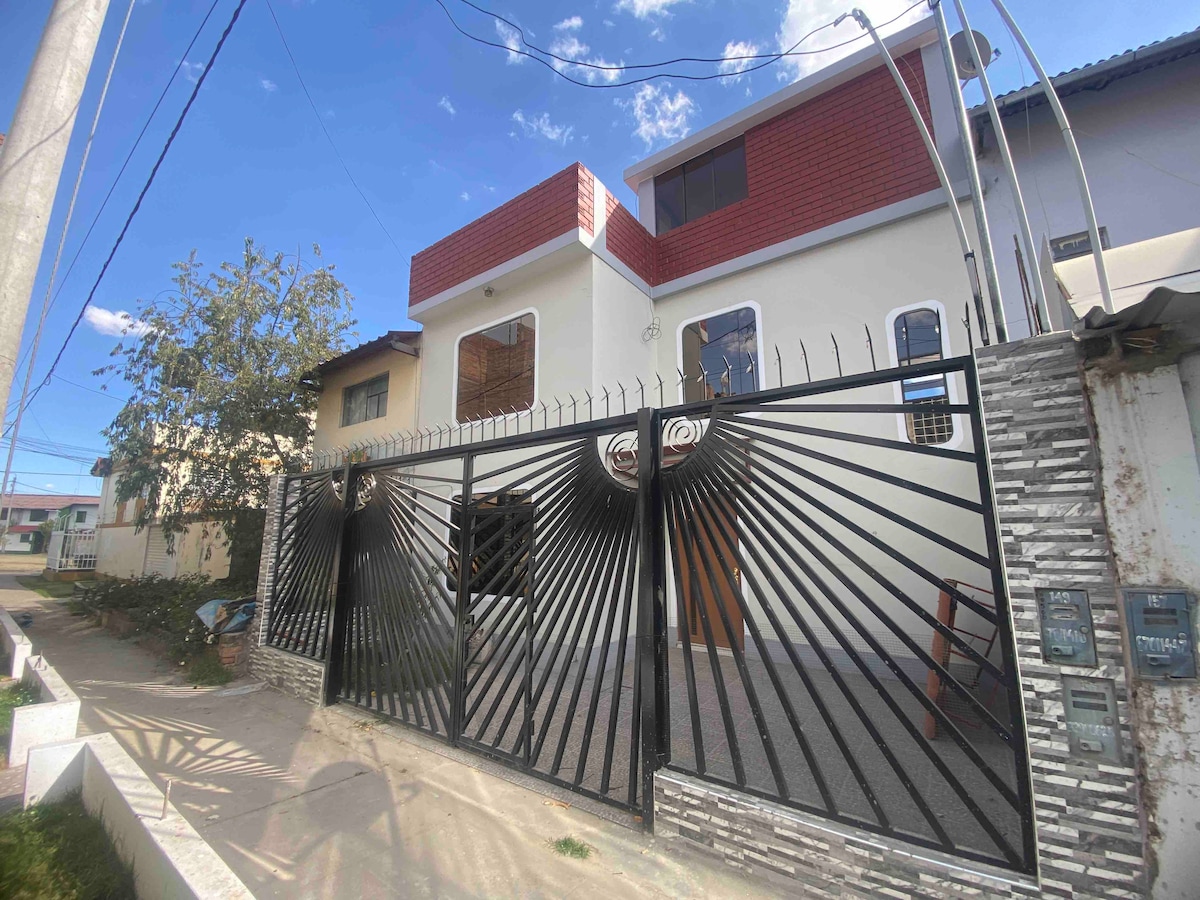 The exterior of a three-story house is displayed, featuring a modern design with a striking gate showcasing a geometric pattern. The entrance is highlighted by large windows that provide natural light. A clear blue sky complements the facade, surrounded by greenery.