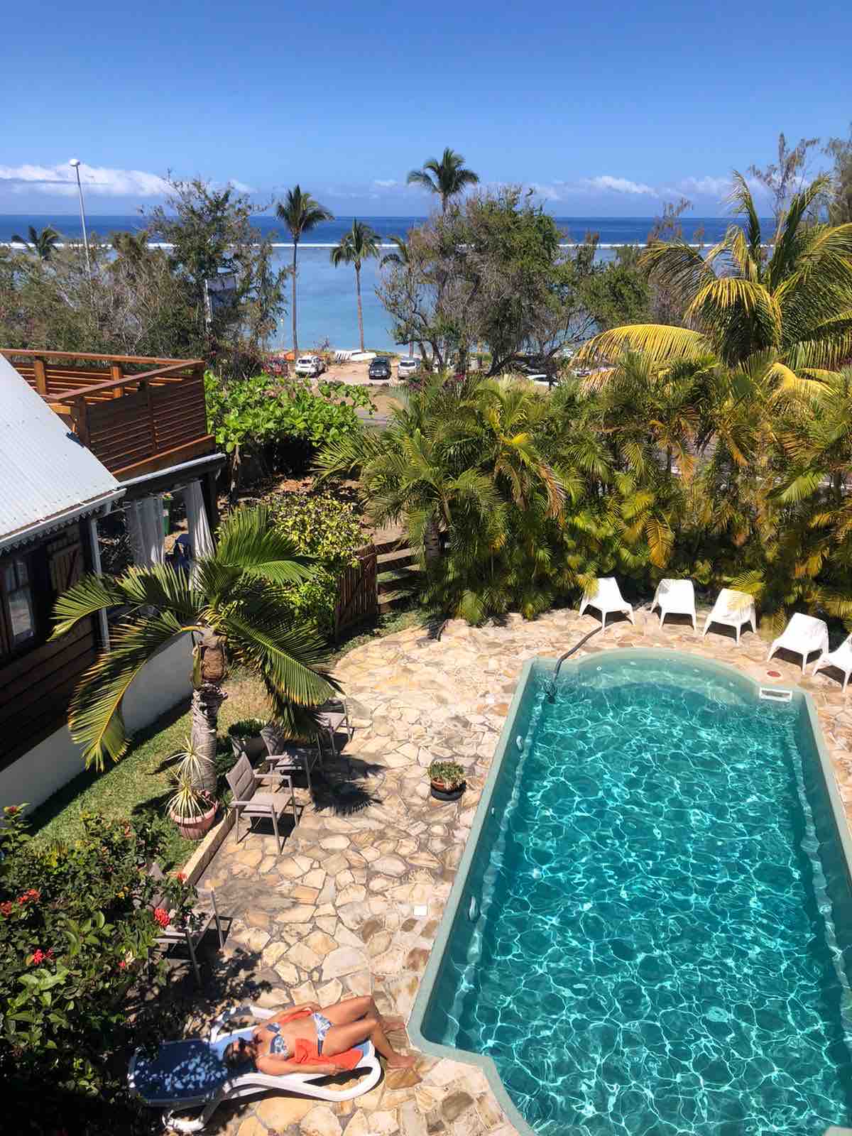 A clear view of a shared swimming pool surrounded by lush palm trees and tropical plants. Several sun loungers are positioned along the poolside. In the background, glimpses of the lagoon can be seen, providing a vibrant blue contrast to the greenery.