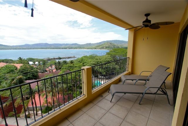 A spacious balcony features two lounge chairs positioned for enjoying panoramic views of Potrero Bay. Sunlight bathes the space, with a ceiling fan providing a gentle breeze. The surrounding greenery and hillside create a calming backdrop, enhancing the relaxing atmosphere.