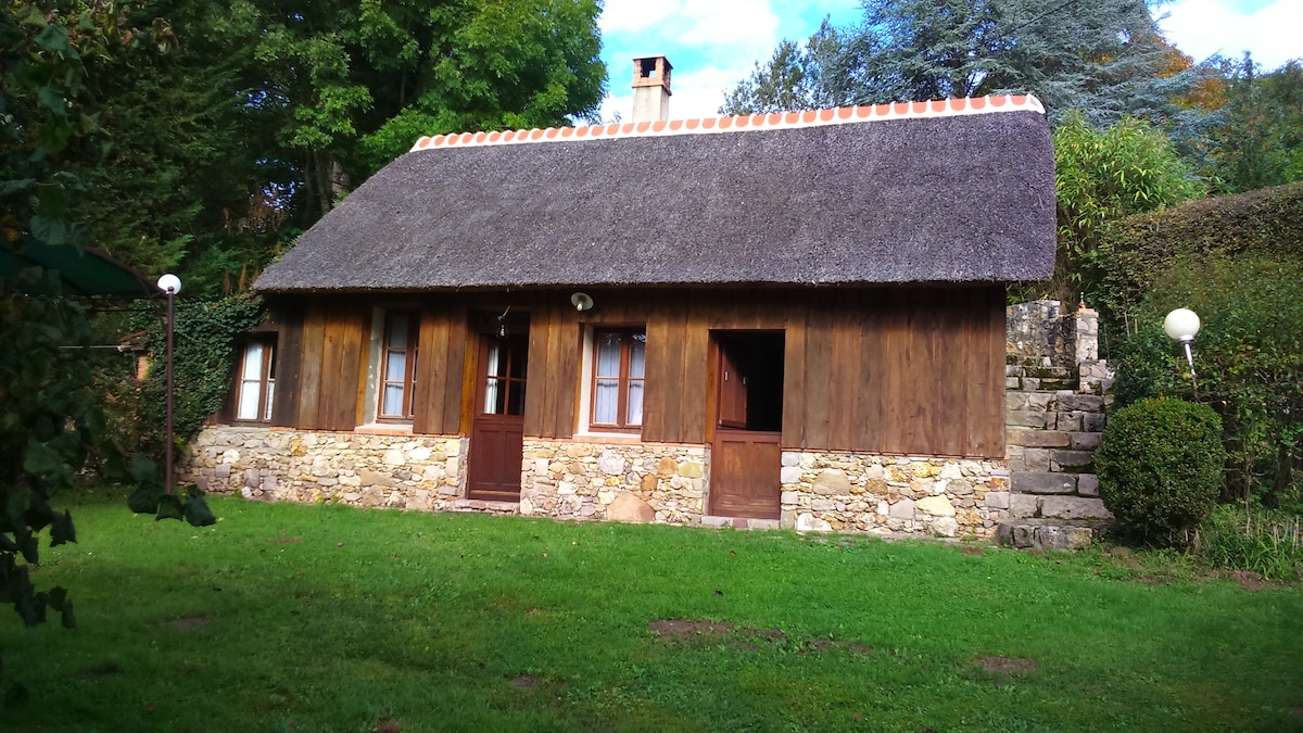 A rustic cottage with a thatched roof is nestled in a grassy area surrounded by trees. The wooden structure features two entrance doors framed by stone walls, complemented by a stone staircase leading up to the property.