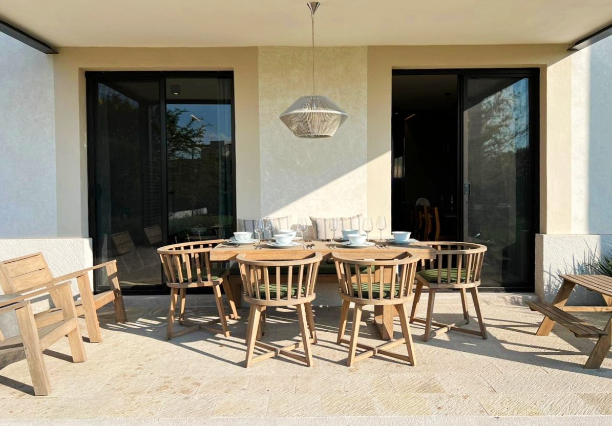 An outdoor dining area features a round wooden table surrounded by eight matching chairs. A contemporary pendant light hangs above the table, and large glass doors lead to the interior spaces. Sunlight casts shadows on the stone flooring, enhancing the setting.
