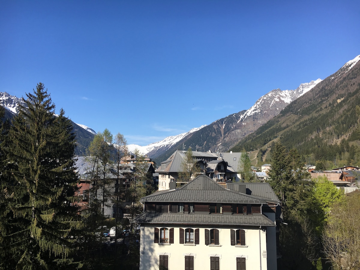 A panoramic view captures the surrounding mountains and valley, with snow-capped peaks in the distance. The image includes charming buildings with sloped roofs and trees interspersed among them, creating a serene atmosphere in the countryside.