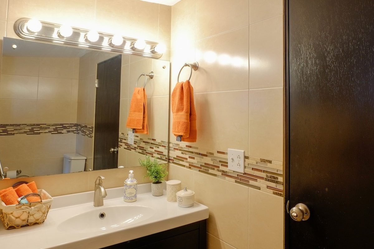 A modern bathroom features a well-lit mirror above a clean countertop. Two orange towels hang on chrome fixtures, while a small plant adds a touch of greenery. Decorative containers and candles sit arranged neatly, enhancing the room's functional elegance.