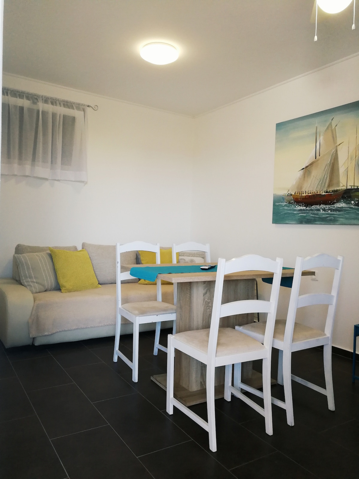 An open-plan dining area is featured, complete with a wooden table surrounded by four white chairs. A light-colored sofa with decorative pillows adds comfort to the space, while a painting of a sailing ship adorns the wall, contributing to the room's coastal theme.