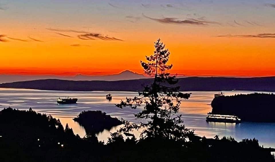 A scenic view captures the vibrant colors of sunset reflected over calm waters. Silhouettes of trees and boats contribute to the tranquil landscape, while layers of mountains create a stunning backdrop against the illuminated sky.