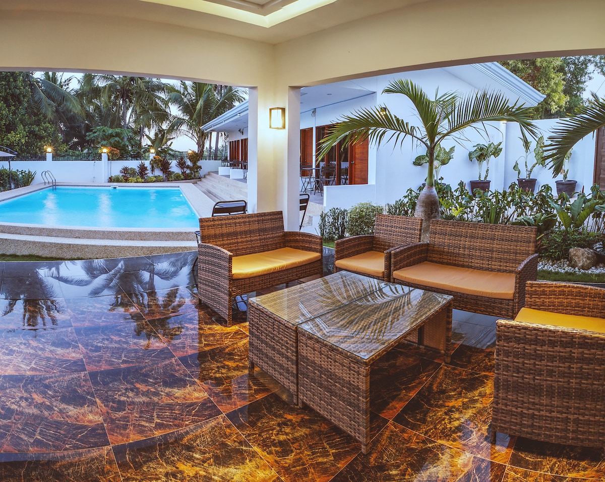 An outdoor seating area features woven furniture with comfortable cushions, arranged around a glass-top table. The space overlooks a swimming pool, surrounded by tropical greenery. A soft glow from a wall-mounted light adds a gentle illumination to the setting.