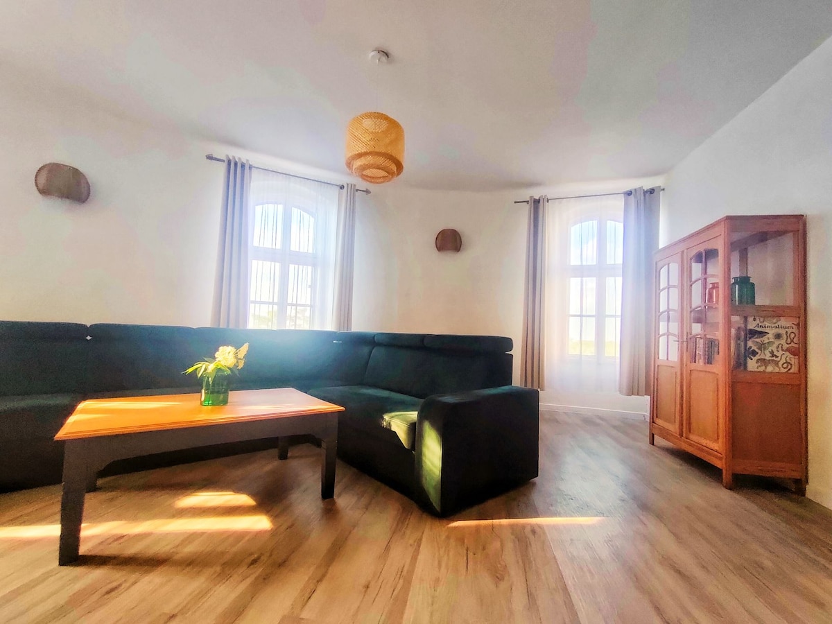 A spacious living area is highlighted by a large dark-colored sectional sofa and a wooden coffee table adorned with a small vase of flowers. Tall windows allow natural light to brighten the room, while a wooden cabinet provides additional storage.