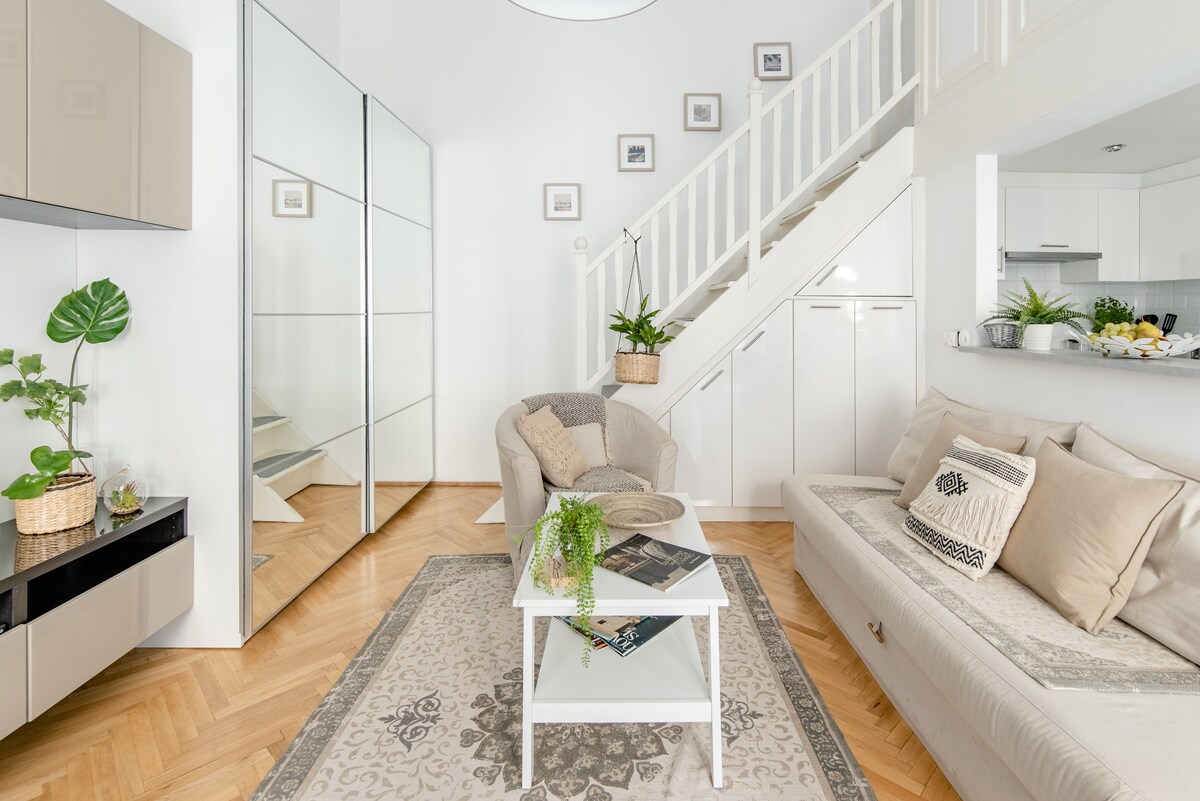 A light-filled living area features a stylish sofa and armchairs positioned around a low table. A large mirror reflects the space, enhancing its openness. Decorative plants add a touch of greenery, while soft textiles and a patterned rug provide comfort underfoot.