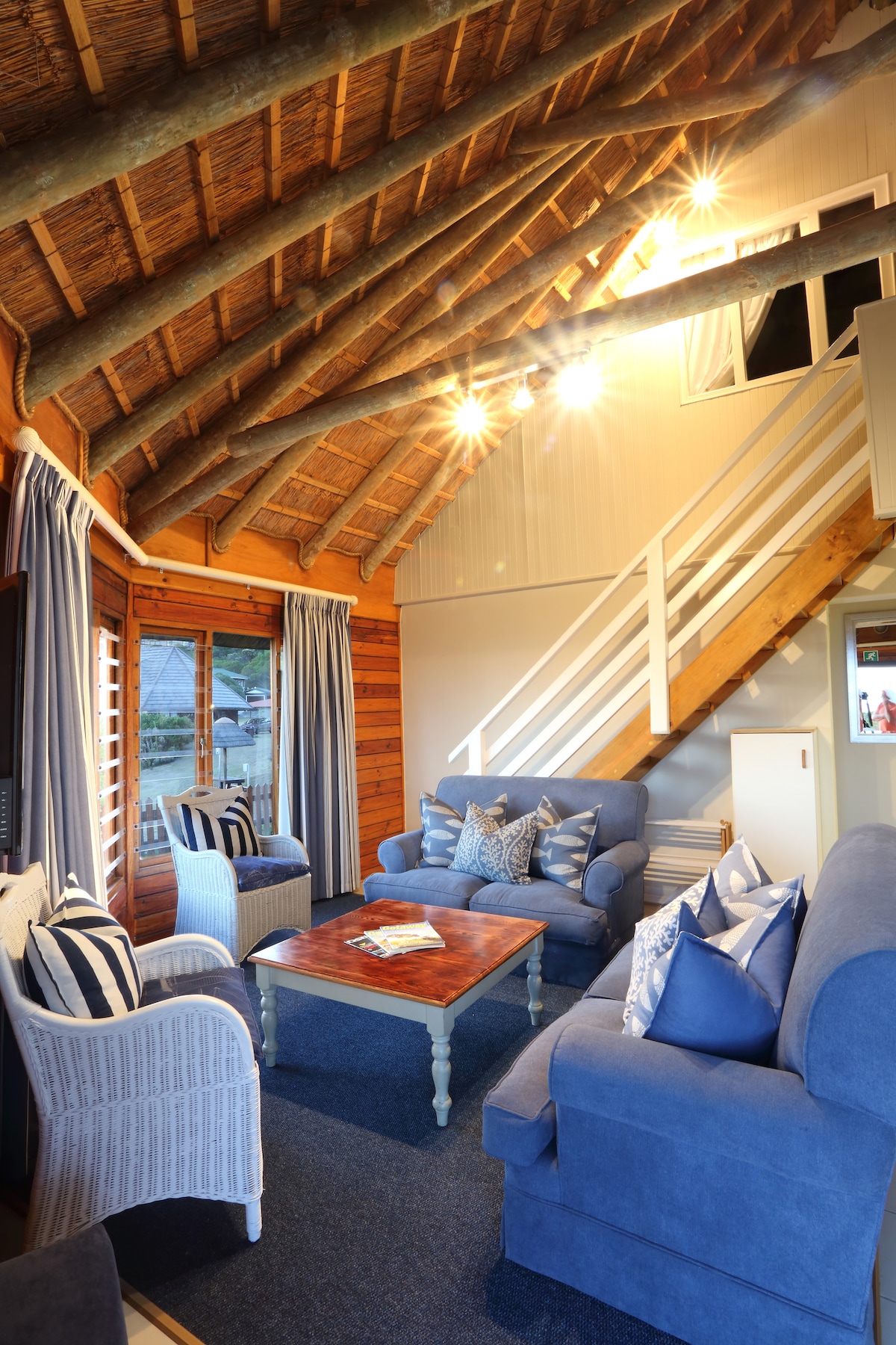 A spacious lounge area features textured wooden walls and a high thatched ceiling. Comfortable blue sofas and wicker chairs surround a wooden coffee table, accented by decorative pillows. Large windows allow natural light to fill the space, creating a cozy atmosphere.