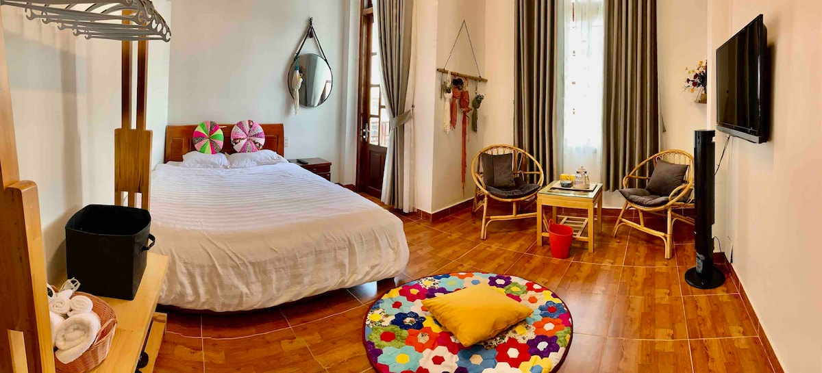 A bright and airy bedroom features a spacious bed with white linens, complemented by a colorful round rug. Two comfortable chairs and a small table are placed near a window, while a mirror and wall decor add to the inviting setting.