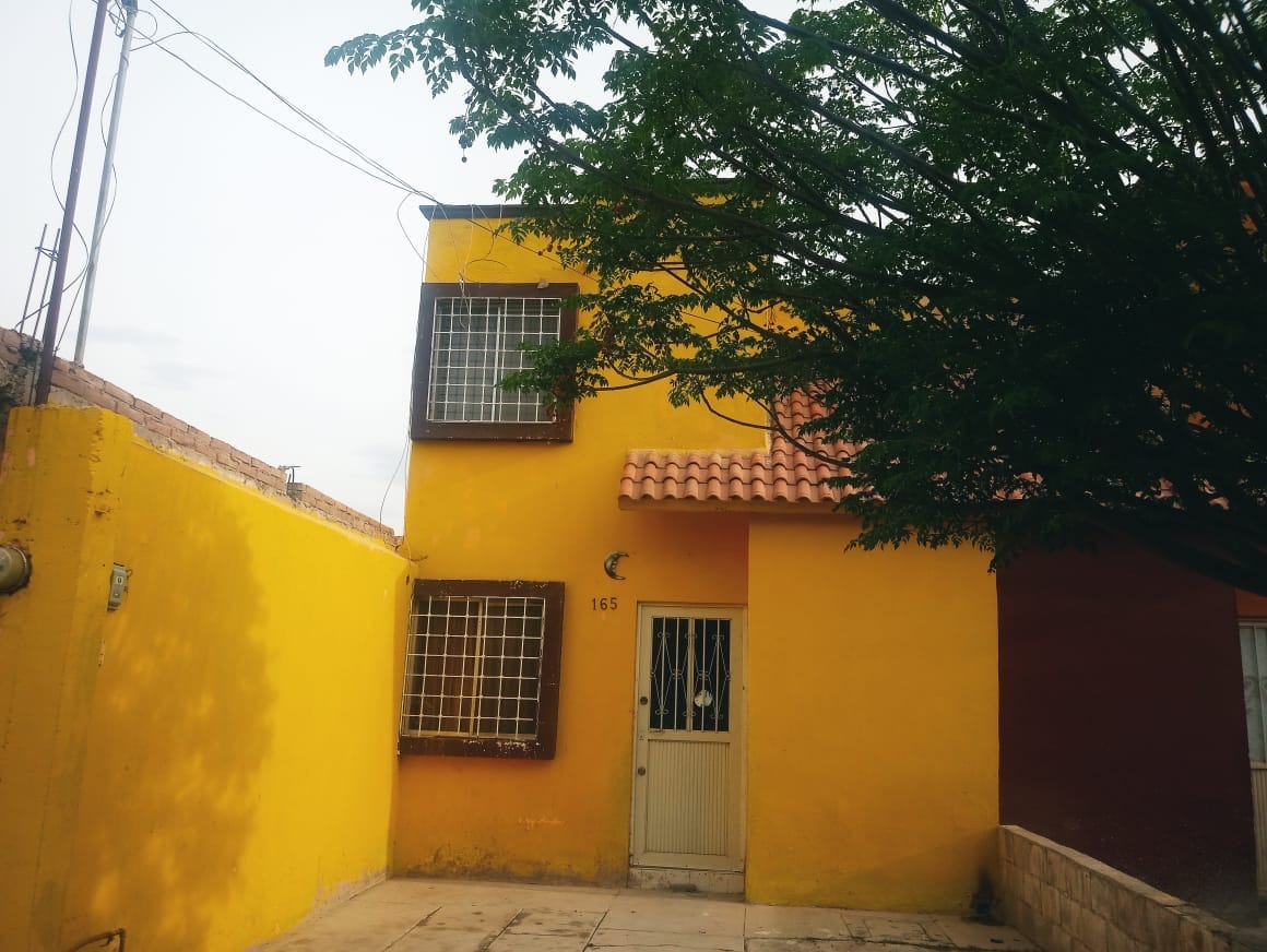 A traditional house exterior features vibrant yellow and red walls, complemented by a terra-cotta tiled roof. Large windows with bars add character, while a lush tree provides shade in the front yard, creating a welcoming atmosphere for relaxation.