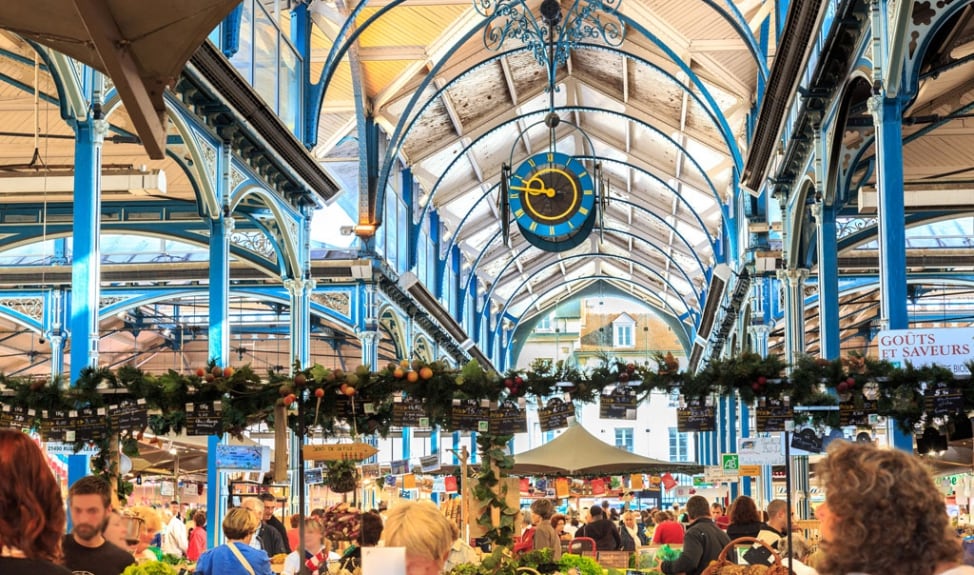 The image portrays a bustling market interior, characterized by high ceilings adorned with intricate blue beams and a prominent clock. Stalls filled with various goods create a lively atmosphere, while a variety of shoppers can be seen engaged in exploration and conversation.