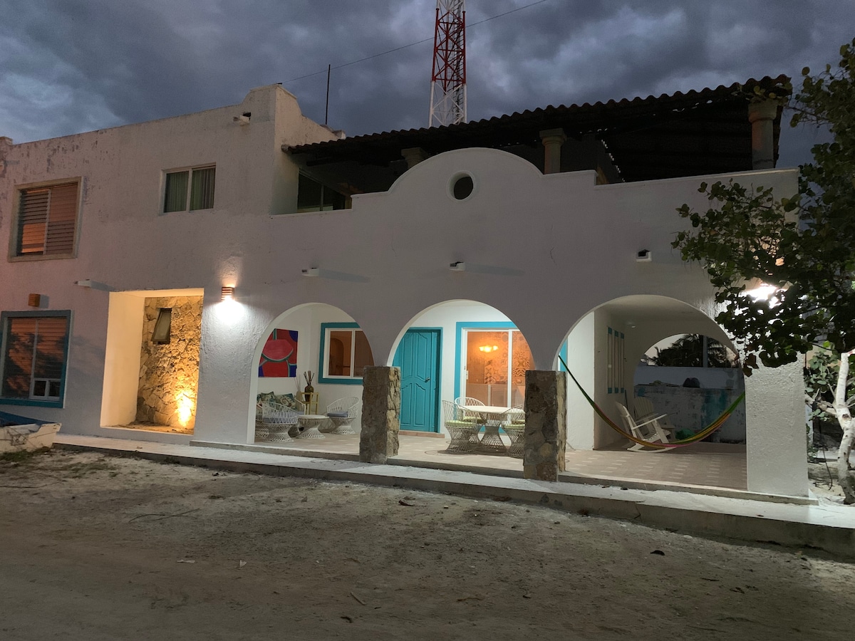 A charming two-story home with a white exterior is showcased under a cloudy sky. The front features arched doorways and a spacious patio furnished with woven chairs and a hammock, inviting relaxation. Soft lighting enhances the architectural details, creating a welcoming entryway.