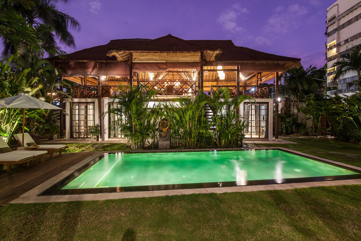 A spacious villa is set against a twilight sky, displaying a private swimming pool with inviting green water. Lush tropical greenery surrounds the area, and the villa's large windows reflect warm light from within, enhancing a serene outdoor ambiance.
