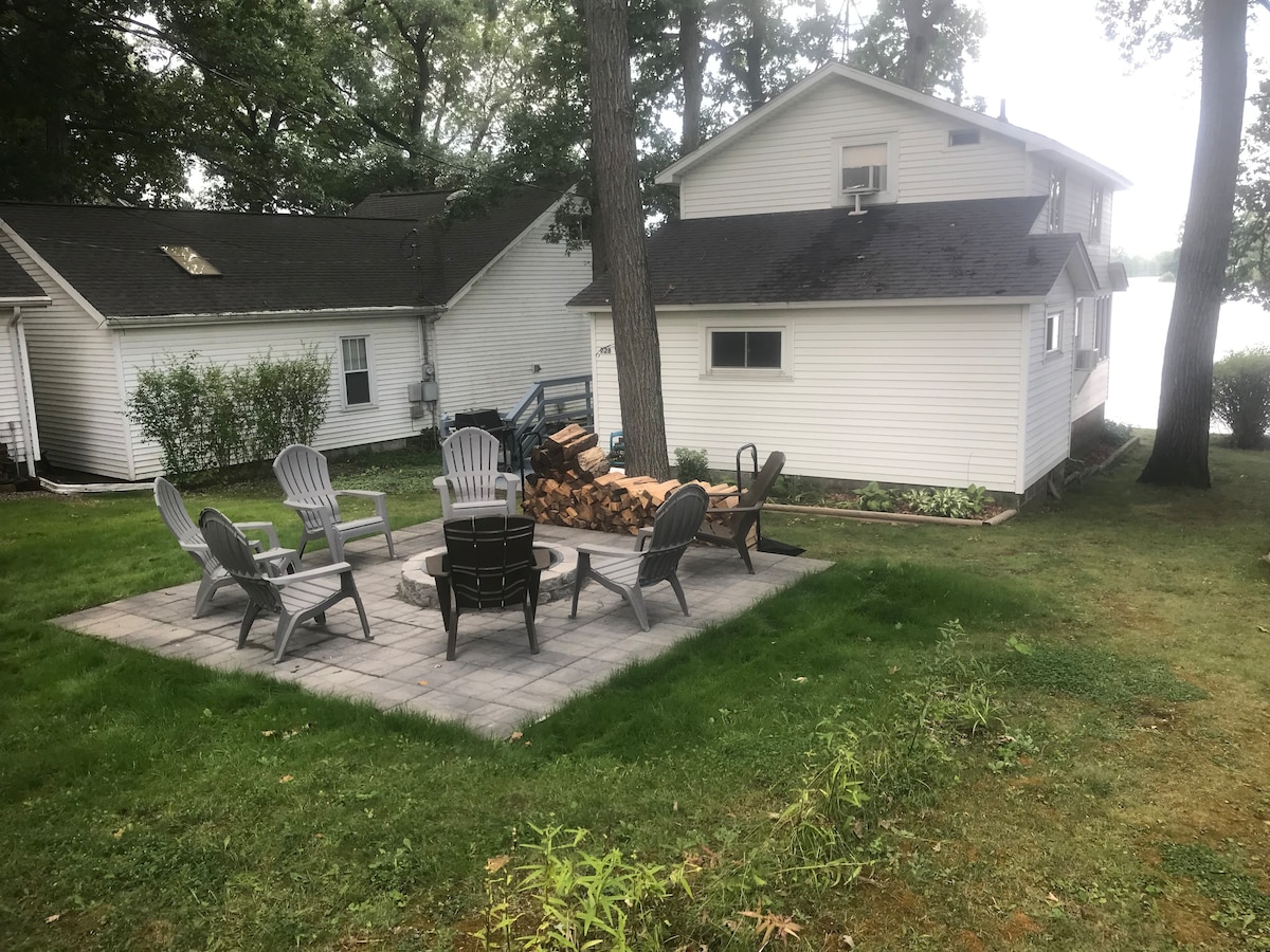 A cozy outdoor seating area features six Adirondack chairs arranged around a circular stone patio, with a fire pit at the center. A stack of firewood is positioned nearby, and the scenic lake is visible in the background, framed by trees.