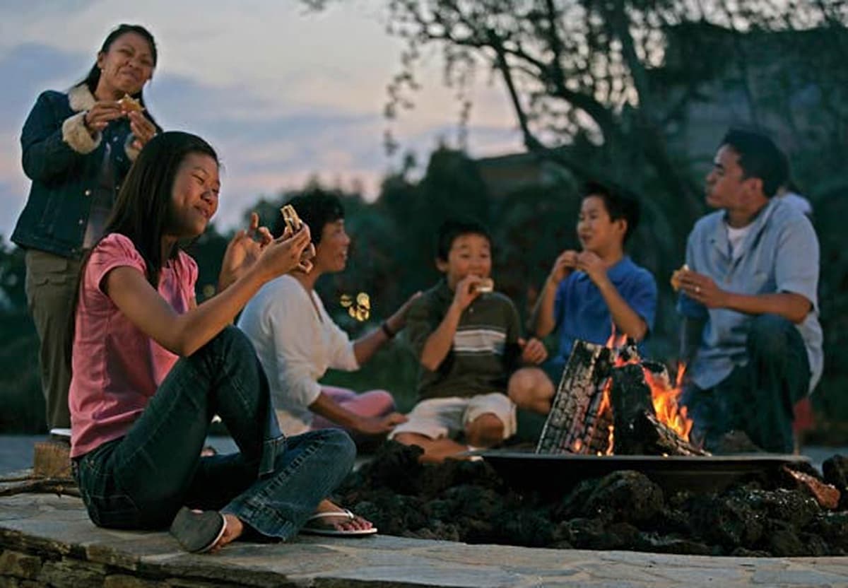 A gathering around a fire pit is depicted, with individuals seated on a stone perimeter. The flames flicker brightly, surrounded by snacks and casual clothing. The scene is complemented by a tranquil sunset backdrop, creating a relaxed atmosphere for socializing.