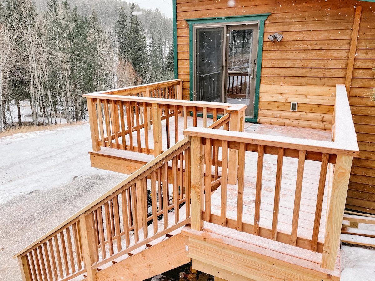 A private deck made of natural wood is showcased, featuring railings and wide steps leading down. The deck is covered in a light dusting of snow, with a view of surrounding trees and the lightly snow-covered landscape.
