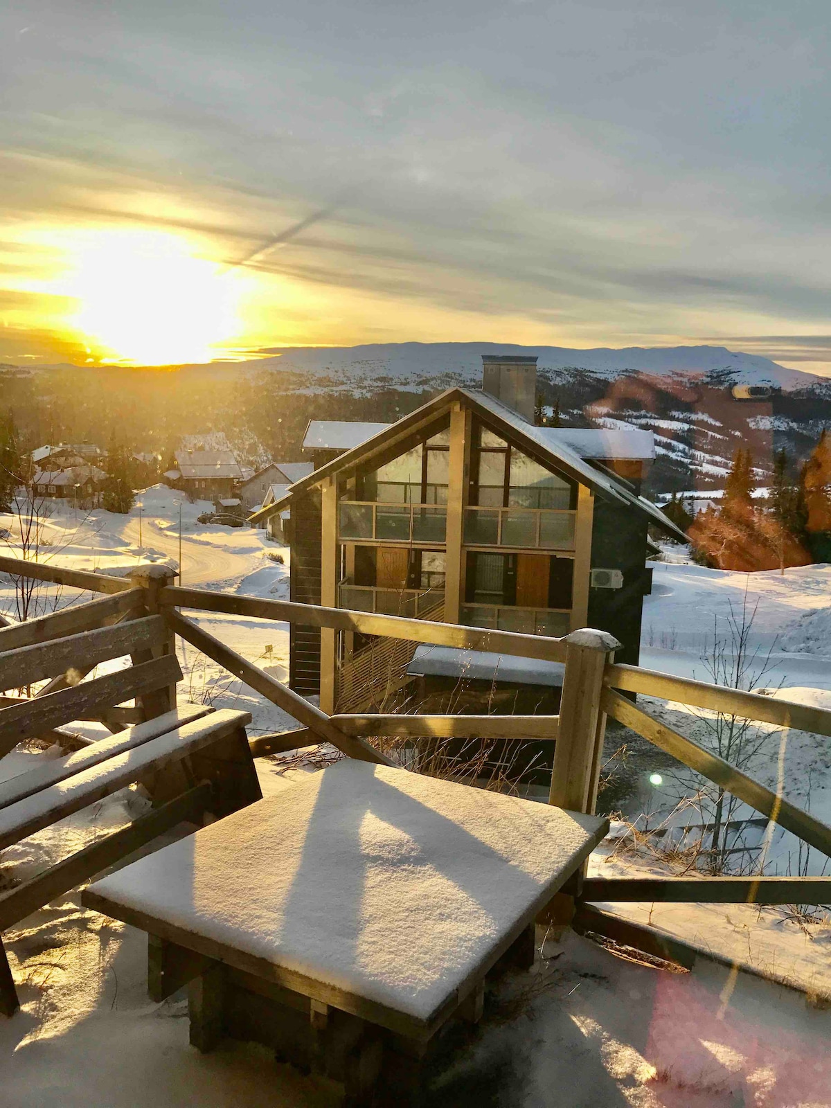 A winter scene showcases a beautiful building with large windows, framed by snow-covered terrain. The sunrise casts a warm glow over the landscape, illuminating distant mountains and creating a peaceful atmosphere. A wooden table is visible, lightly dusted with snow.