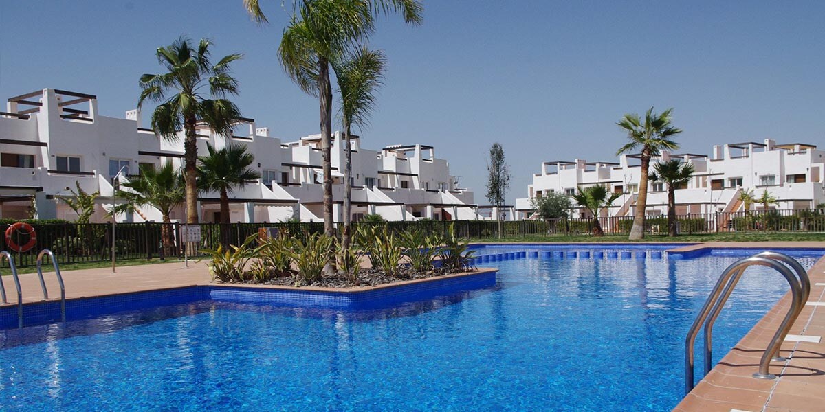 A large communal pool is surrounded by landscaped gardens. The water reflects the clear blue sky, while palm trees and shrubs add greenery to the scene. Modern white buildings with balconies and terraces are visible in the background, enhancing the resort's inviting atmosphere.