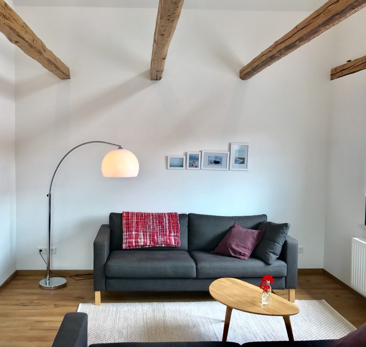 A spacious living area is highlighted by wooden beams and natural light. A dark sofa is adorned with decorative pillows and a throw blanket. A curved floor lamp stands nearby, illuminating a small round table with a flower vase, complemented by artwork on the wall.