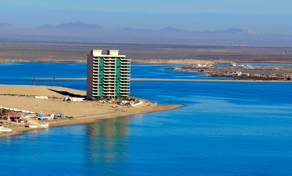 A distant view of a modern high-rise building situated along a serene coastline, surrounded by calm, azure waters. The beachfront area is lined with sandy shores, while the horizon features distant mountains and a clear sky, emphasizing the tranquil landscape.