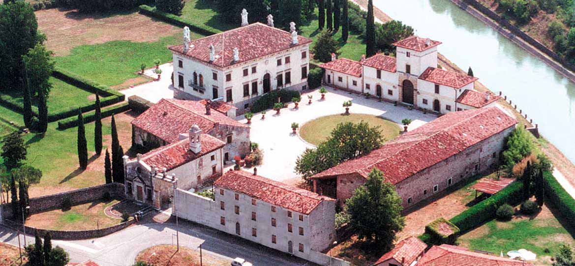 The aerial view showcases the expansive grounds of Villa d'Arco, featuring beautifully manicured gardens, quaint pathways, and characteristic terracotta roofs. Surrounding greenery complements the timeless architecture of the buildings, creating a harmonious environment that embodies historical elegance.