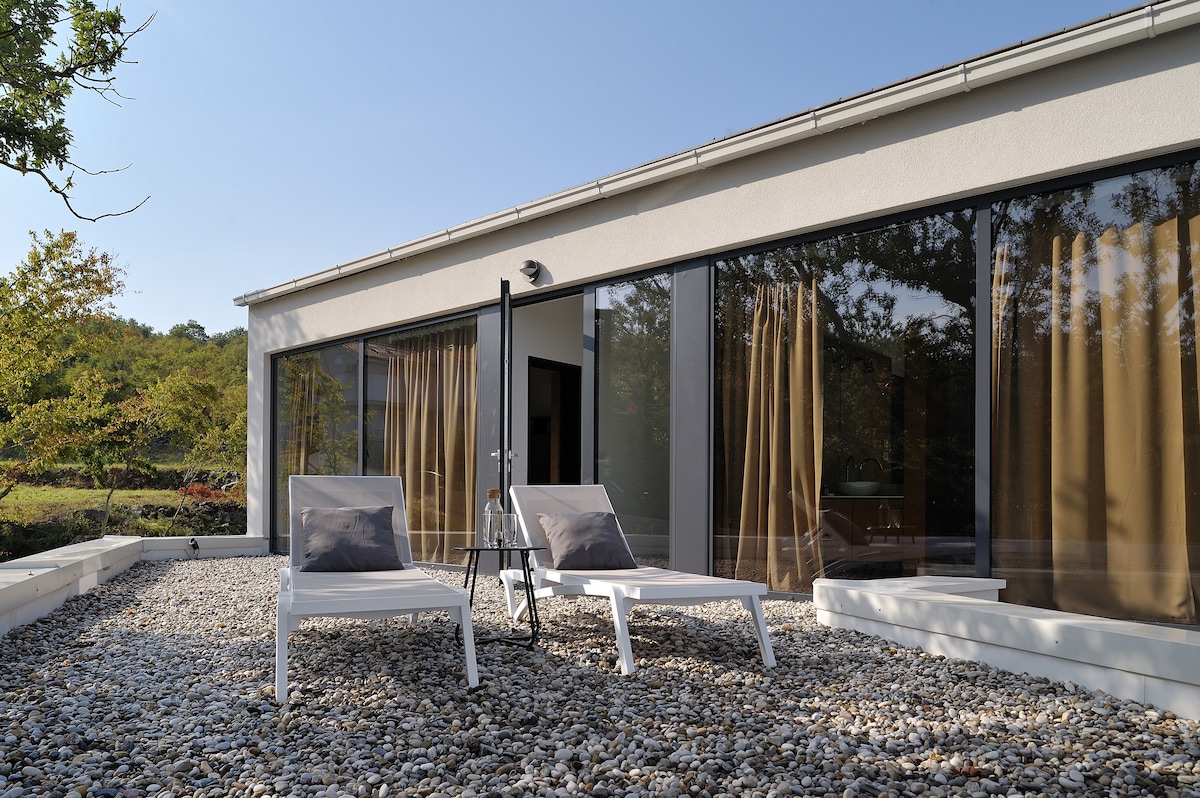 Two loungers are positioned on a pebble terrace, with soft cushions adding comfort. Large glass doors provide a view into the living space, and trees can be seen in the background, enhancing the connection to nature.