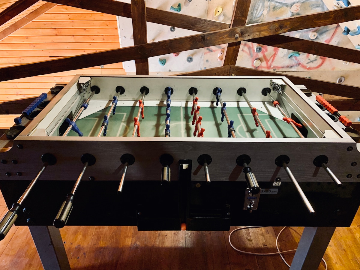 A foosball table is positioned in a spacious game area, featuring colorful players in red and blue. Wooden beams create an open, rustic ambiance above the table, enhancing the recreational setting.
