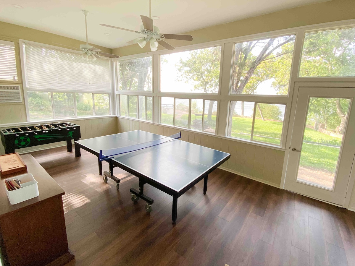 The sunlit game room features two large tables designed for playing ping pong, surrounded by expansive windows that provide a view of the outdoor greenery. The flooring is dark wood, complemented by a spacious door leading to the outside.