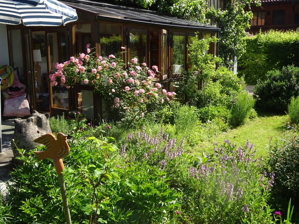 A landscaped garden adorned with vibrant flowers in various shades. The area features a lush green lawn and blooming rose bushes, alongside a sunlit terrace area with large windows, providing a view into the adjacent living space.