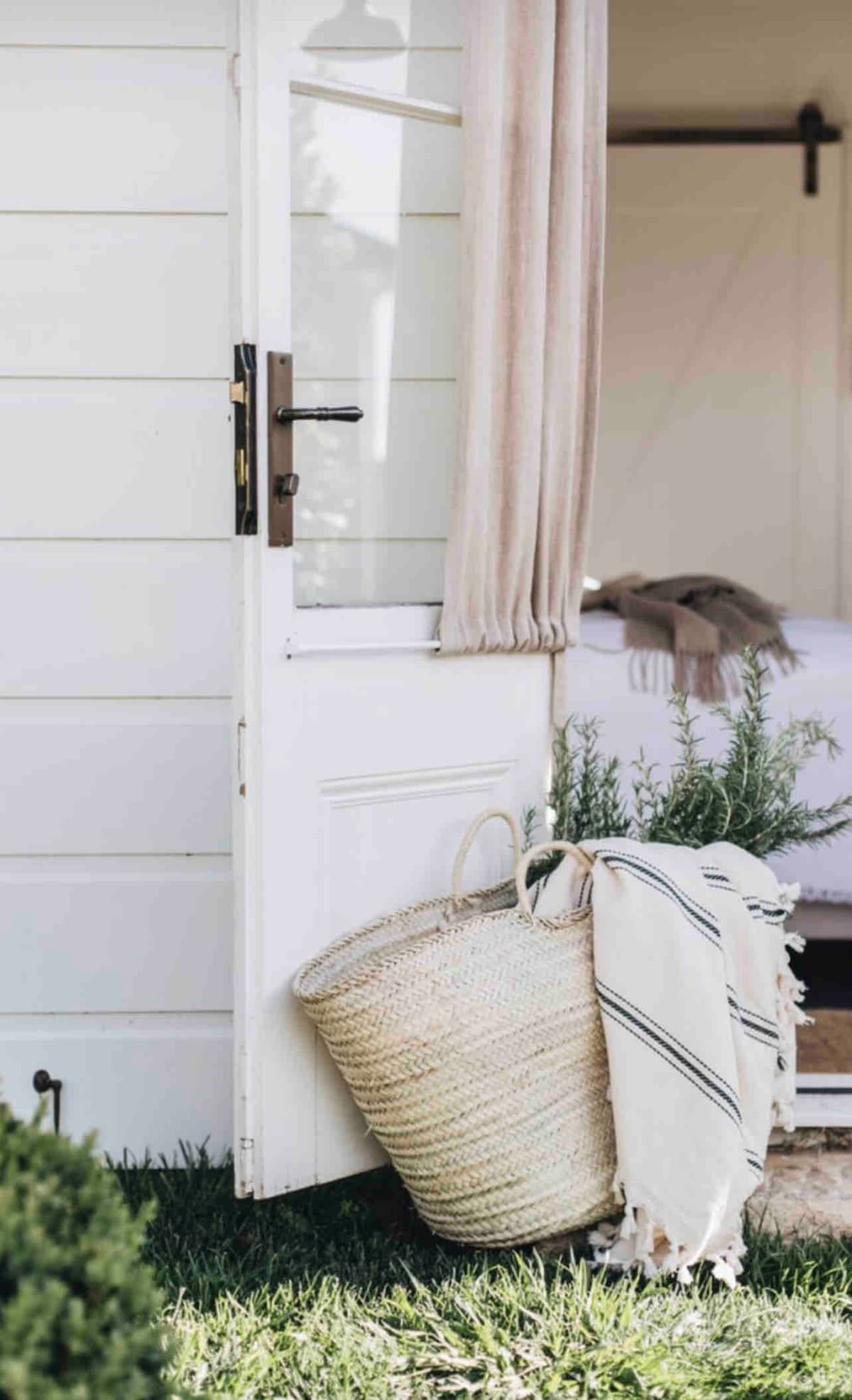 An open door leads into a guest room, framed by softly draped curtains. A woven basket rests on the grass, featuring a striped textile. Lush greenery surrounds the doorway, complementing the inviting entryway.