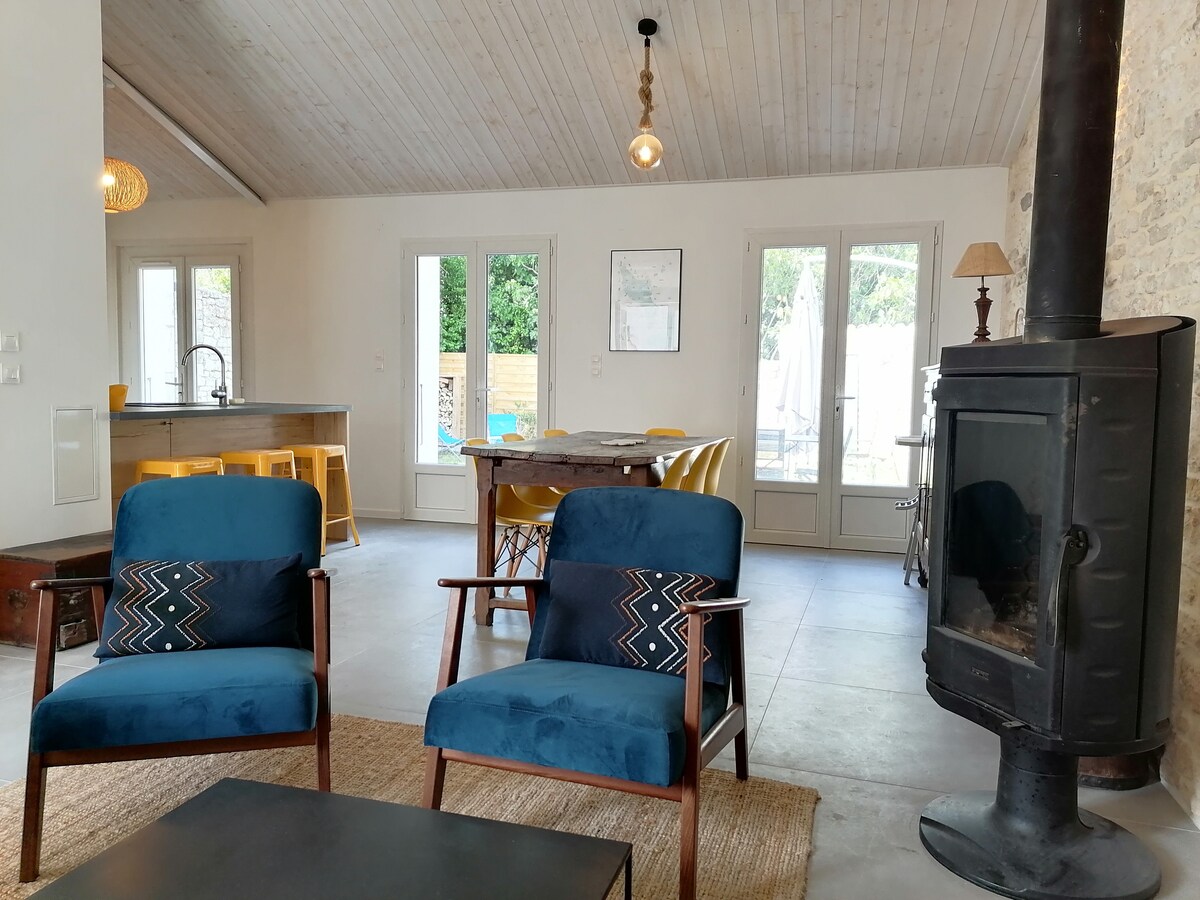 A cozy living area features two blue armchairs with decorative cushions positioned near a wood stove. An open layout reveals a dining table surrounded by yellow chairs, leading to large glass doors that offer views of the patio.