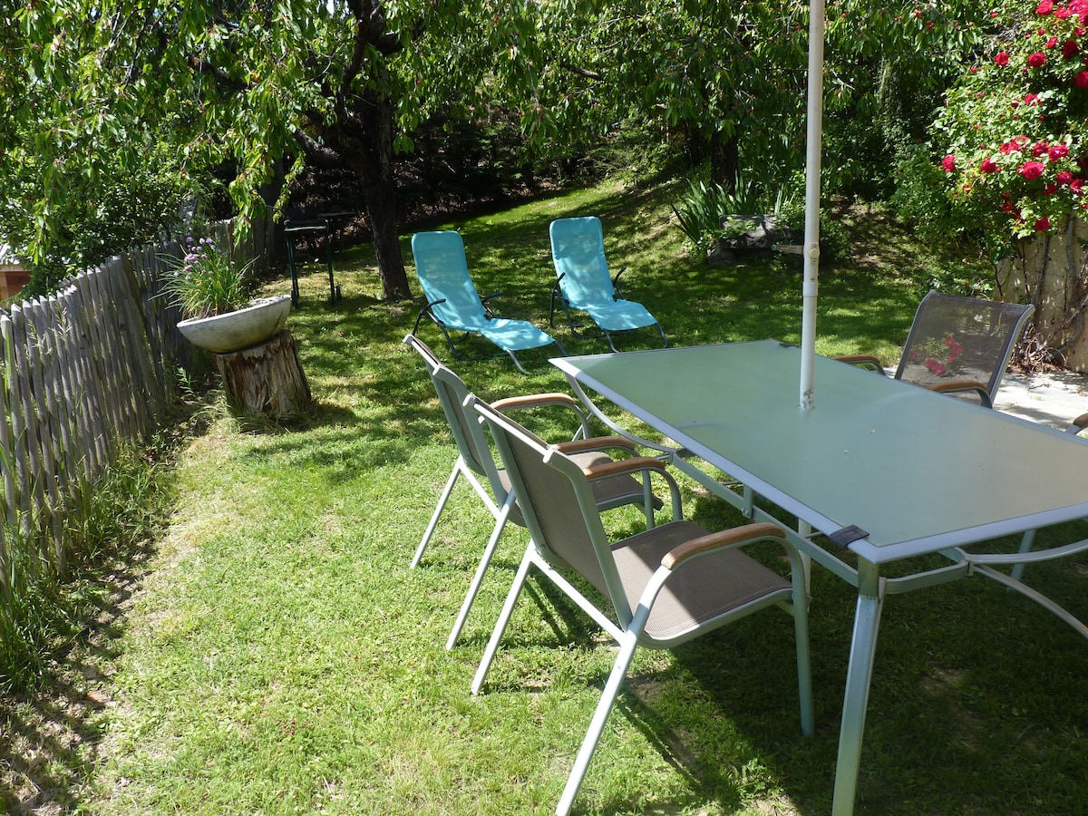 A spacious garden area is depicted, featuring a dining table and several chairs arranged for outdoor enjoyment. Two lounge chairs are positioned under the shade of green foliage, while a decorative bowl and vibrant flowers add visual interest to the landscape.