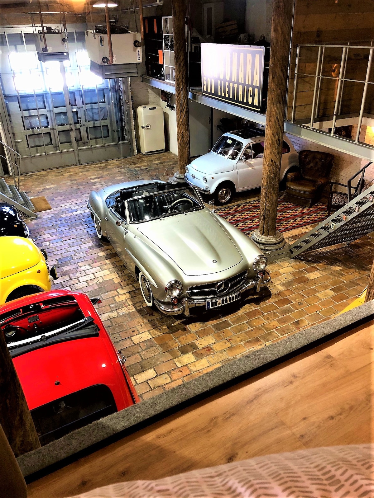 A vintage silver convertible is parked among several classic cars in a spacious garage, showcasing a mix of colors and styles. The floor is paved with decorative stones, and large windows illuminate the area, enhancing the industrial feel of the setting.