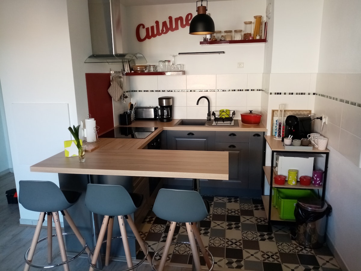 The kitchen area features modern appliances and ample counter space. A central island with three bar stools provides seating. Open shelving displays kitchenware and decorative items. The tiled floor adds a stylish touch, complemented by natural light coming from a window.