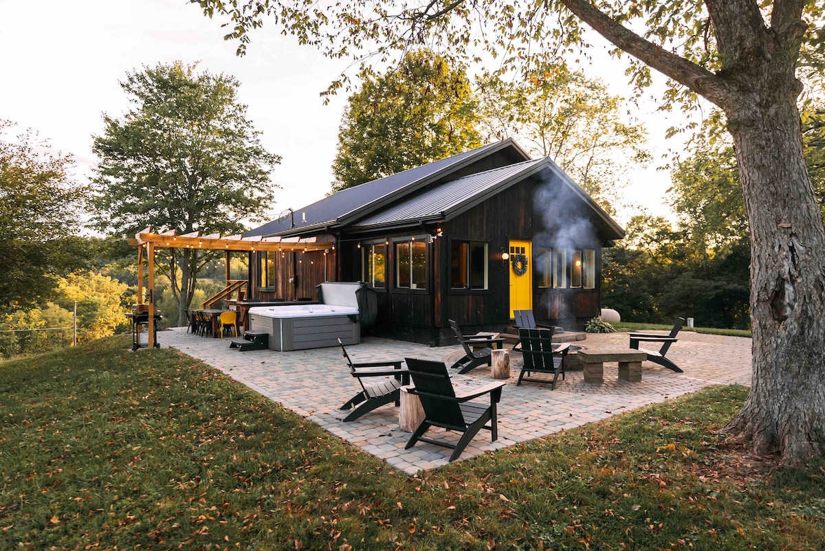 A rustic cabin exterior is set against lush greenery, featuring a stone patio with seating around a fire pit. A hot tub is visible on the deck, and a yellow door adds a bright accent to the dark wood façade.
