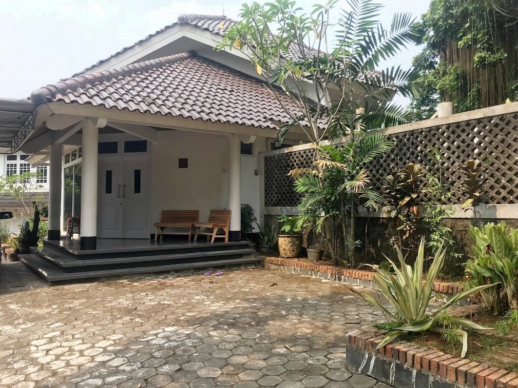 The exterior of the villa is presented with a traditional roof and a welcoming porch, featuring two wooden benches. Tropical plants and greenery frame the entrance, while a stone pathway adds to the charm. The surrounding area includes low brick walls and potted plants.