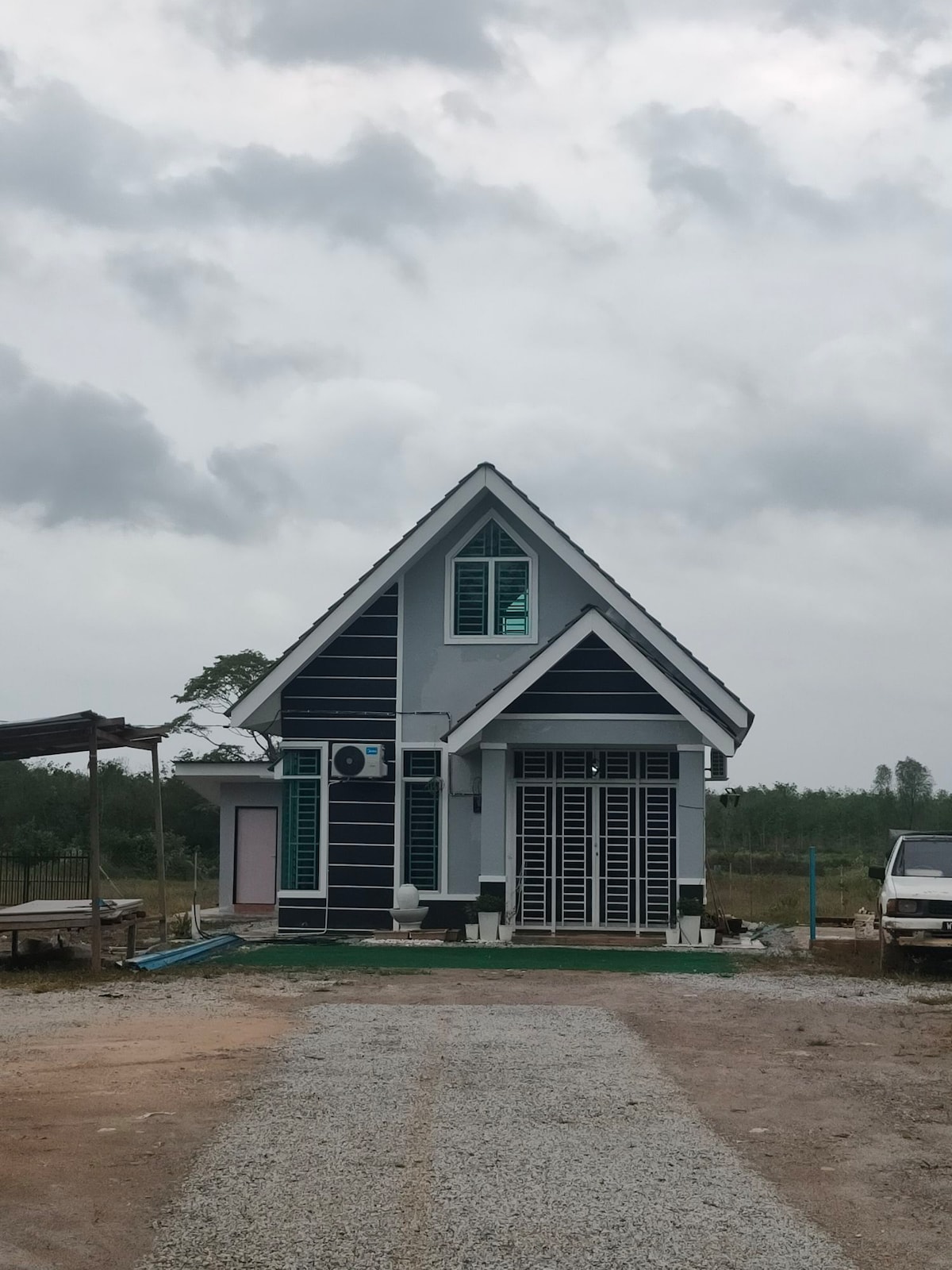 A modern house features a pitched roof and blue exterior accents. Large windows provide natural light and ventilation. A small porch with sliding doors is visible, along with an air conditioning unit mounted on the wall. The surrounding area consists of gravel and open land.