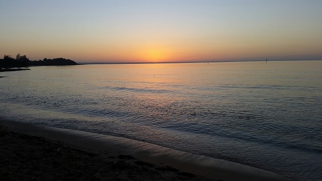 A serene beach scene captures the calm waters reflecting the warm hues of a sunset. The horizon displays a gentle gradient from orange to deep blue, while the shoreline is lined with soft sand. Silhouetted trees create a tranquil backdrop.