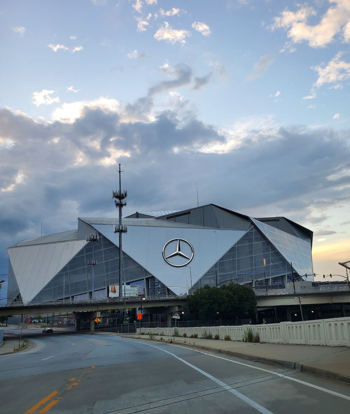 The image shows the modern exterior of Mercedes-Benz Stadium, characterized by its striking geometric design and large glass panels reflecting the sky. The prominent Mercedes-Benz logo is displayed on the front, with surrounding roadways and greenery visible in the foreground.