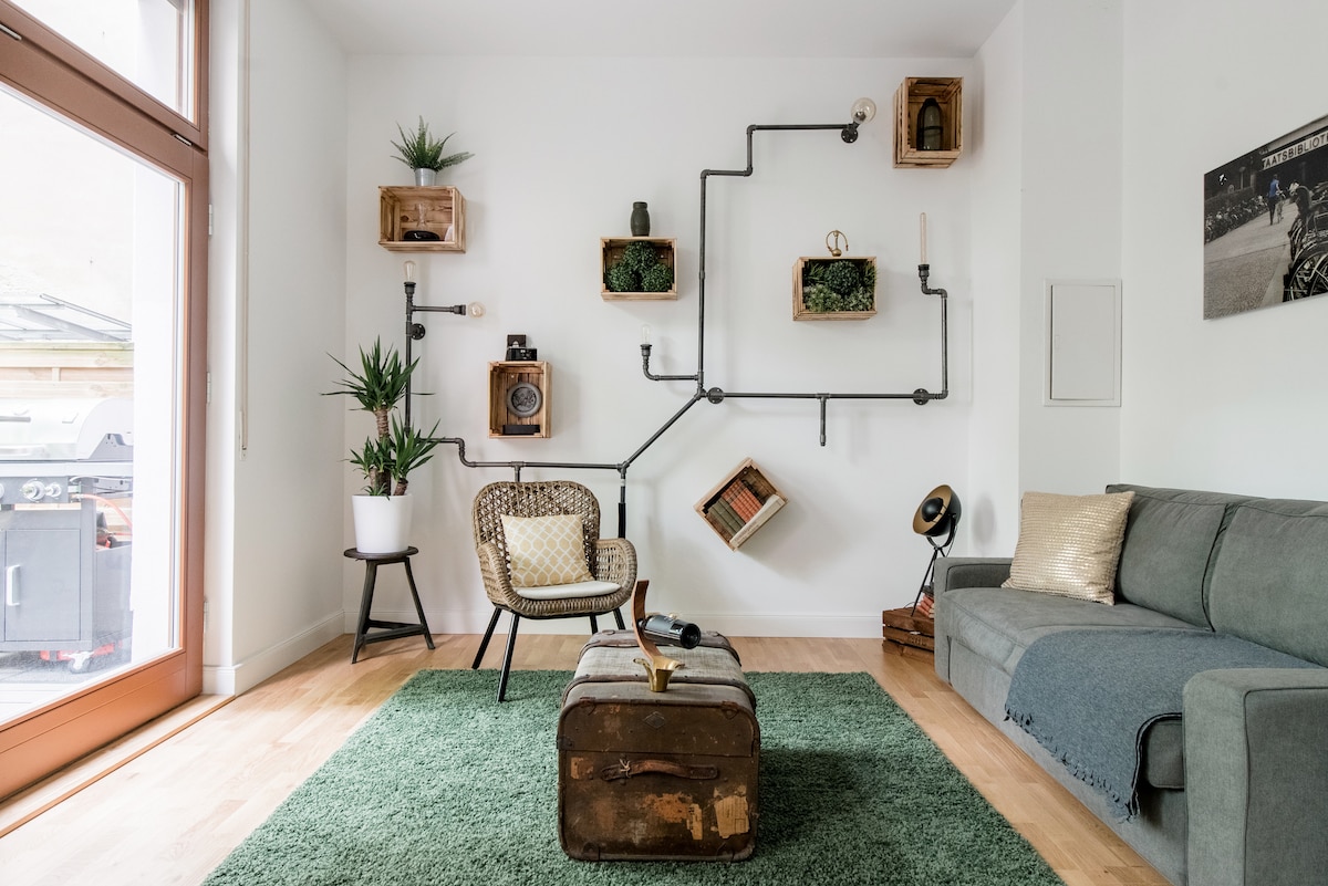 A cozy living area features a light grey sofa and an armchair with a patterned cushion. Green plants add a natural touch, while wall-mounted wooden boxes and piping create an artistic display. A vintage trunk serves as a coffee table on a soft green rug.