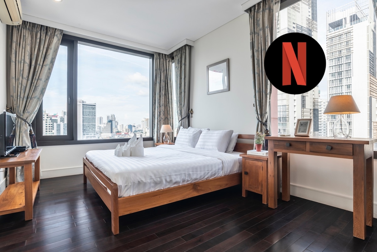 A spacious bedroom features a large bed, wooden furniture, and two windows offering city views. Natural light fills the room, enhanced by light-colored curtains. A television mounted on the wall enables access to streaming services, providing entertainment options during the stay.