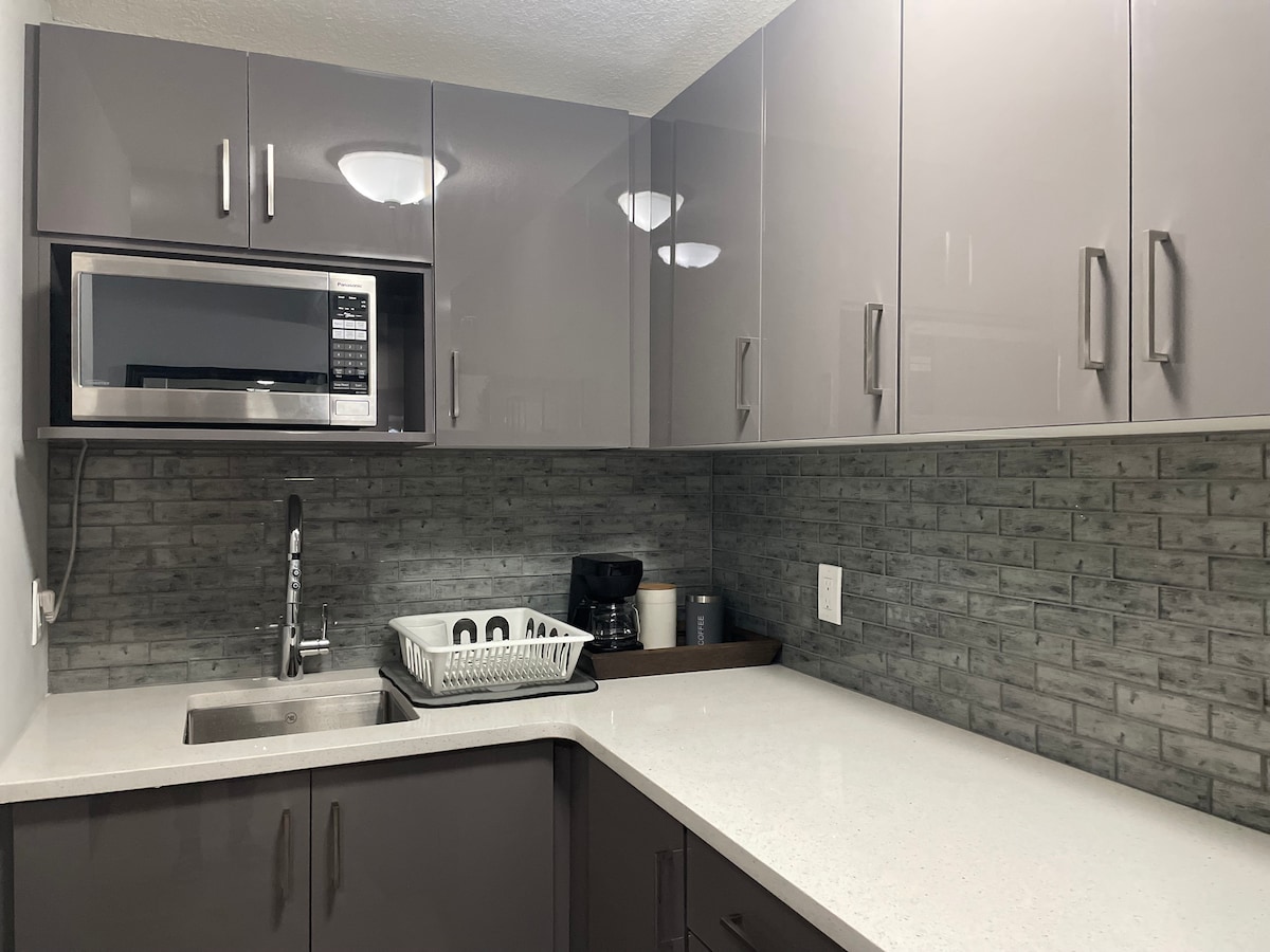 The kitchenette features modern cabinetry with sleek gray finishes. A microwave is situated above a stainless steel sink, and a dish rack is positioned next to it. A coffee maker and various kitchen items are visible on the countertop, complemented by textured backsplash tiles.