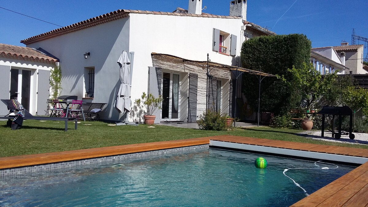 A tranquil garden features a swimming pool with a wooden deck, surrounded by grass. The house, with white walls and tiled roof, displays multiple outdoor seating options including tables and lounge chairs. An umbrella provides shade, while greenery adds a natural touch to the setting.