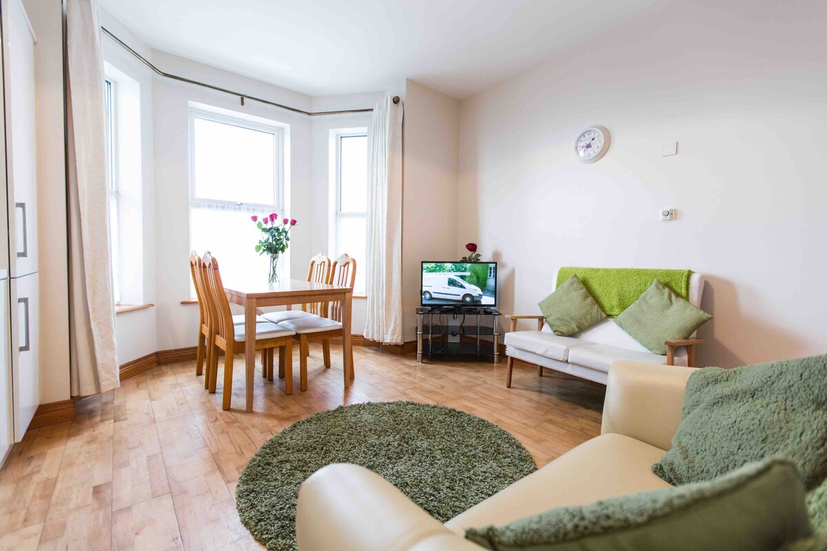 A light-filled living area features a comfortable sofa and a round green rug on a wooden floor. A dining table with wooden chairs is positioned by the windows, adorned with a vase of flowers. A television stands on a low unit, contributing to the cozy setting.