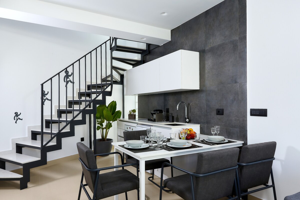 A modern kitchen and dining area are displayed, featuring a sleek white table set with dishware and fruit. Black and grey tones create a contemporary look, complemented by a green plant by the stairs. A minimalist staircase leads to the upper level.