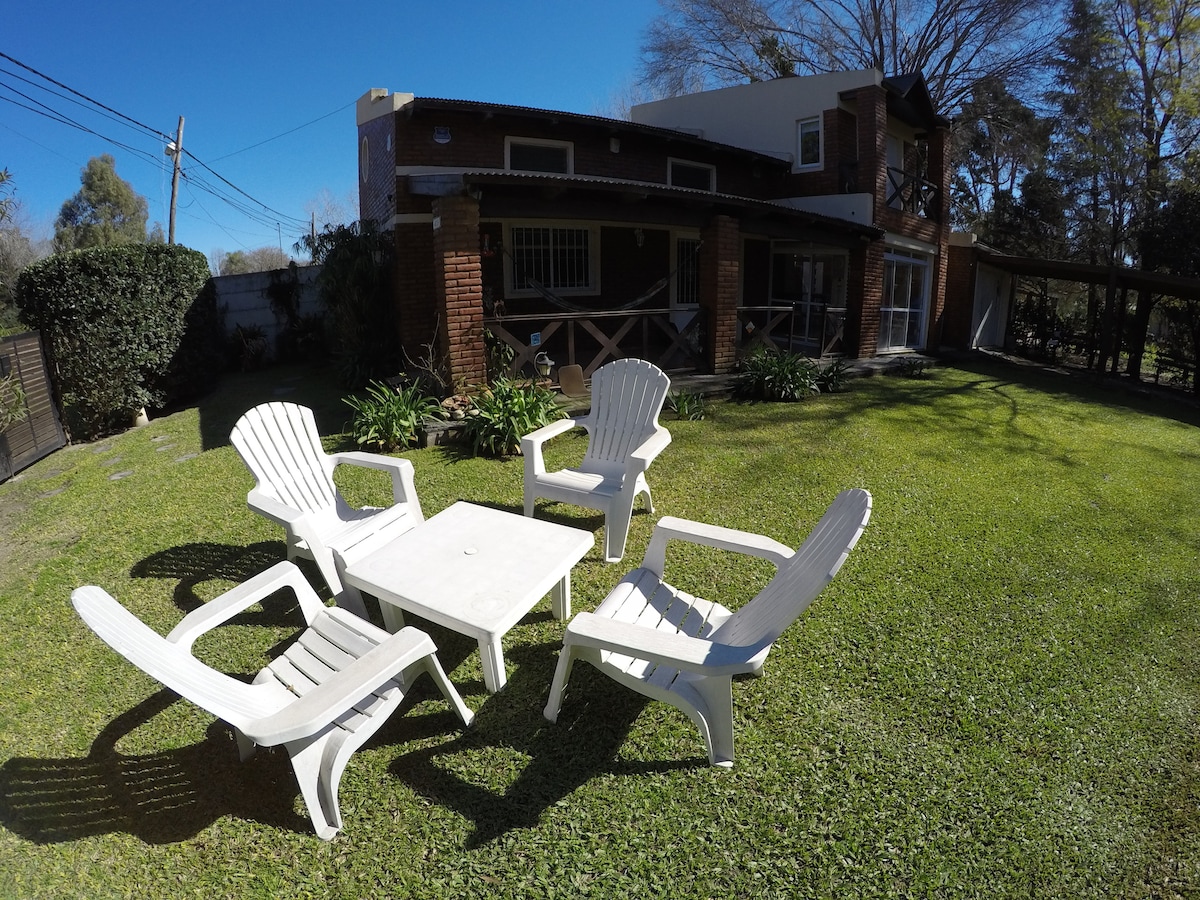 A backyard area is presented with five white chairs arranged around a square table. Lush green grass covers the ground, while the facade of a two-story building is visible in the background, showcasing brick and wooden features. Trees line the property edges.