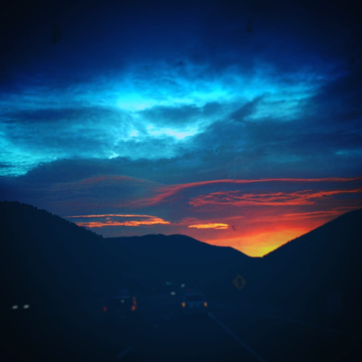The image captures a dramatic sunset over the mountains, with vibrant hues of red and orange contrasting against a deep blue sky. Silhouetted hills frame the scene, while faint outlines of vehicles are visible in the foreground.