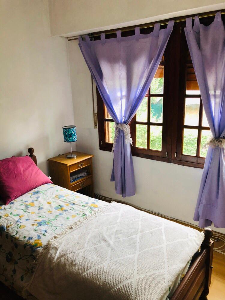 A bright bedroom features a single bed dressed in a floral-patterned blanket. Natural light enters through two wooden-framed windows, adorned with sheer lavender curtains. A bedside table with a decorative lamp adds a functional touch to the warm space.
