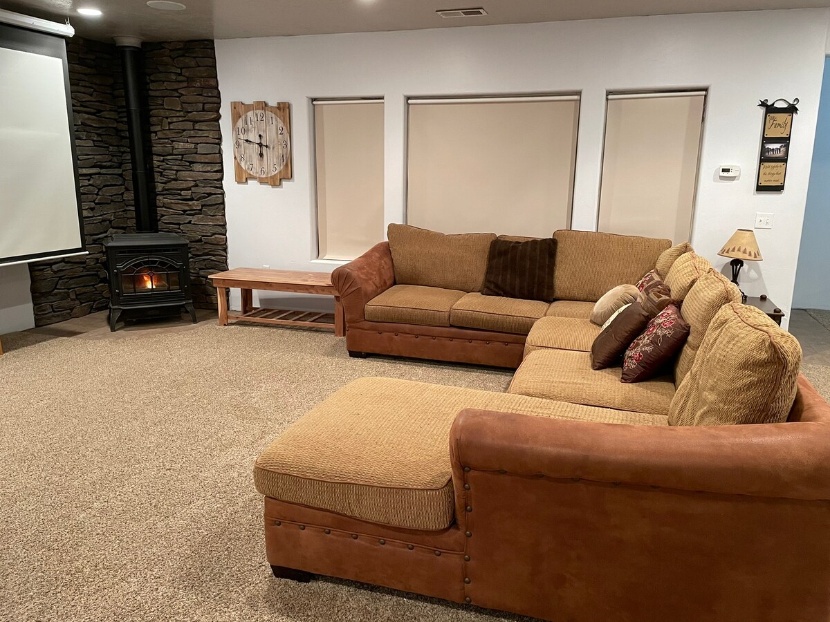 A spacious family room is featured with a large sectional sofa adorned with decorative pillows. A cozy gas fireplace is positioned against a stone wall, beside a flat-screen projector screen. Two windows with shades offer natural light, complementing the warm tones of the carpet.